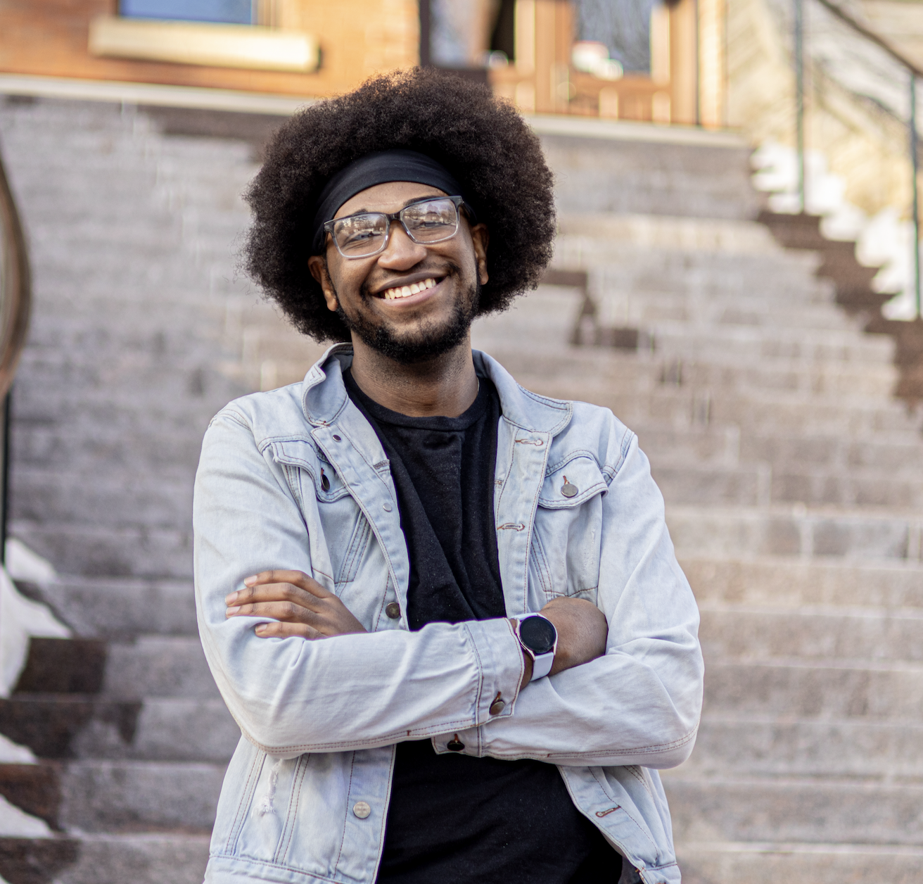 A smiling man with an afro hairstyle and glasses stands confidently with arms crossed in front of a stone staircase. He is wearing a light denim jacket over a black shirt, a black headband, and a smartwatch. Snow is visible on the sides of the steps.